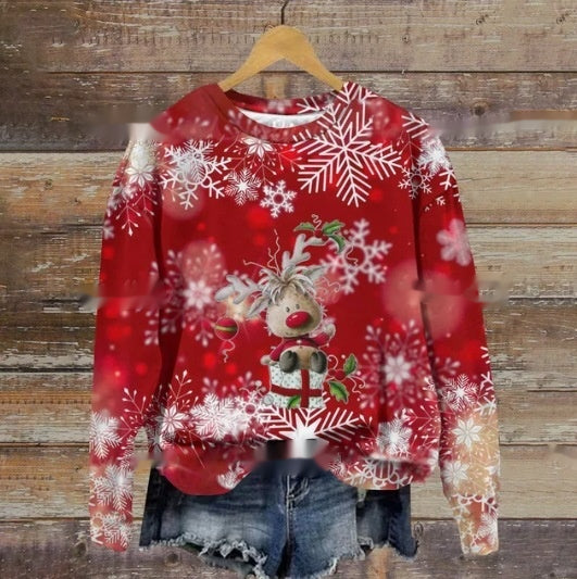 A red Christmas sweater featuring a reindeer with a scarf, surrounded by large white snowflakes, hangs on a wooden background above frayed denim shorts.