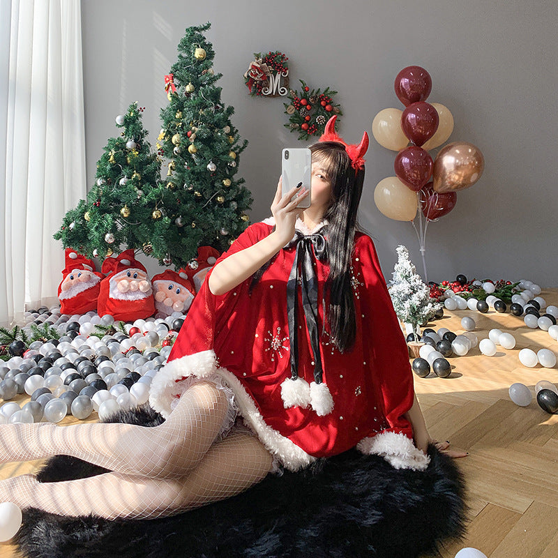 A person in a red festive outfit with devil horns takes a selfie next to Christmas trees and Santa decorations. Balloons and white and black balls decorate the floor.