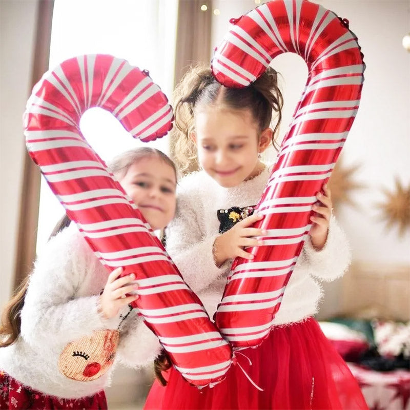 Two children in festive outfits hold large inflatable candy canes, smiling joyfully. The scene is bright and celebratory, with holiday decorations in the background.