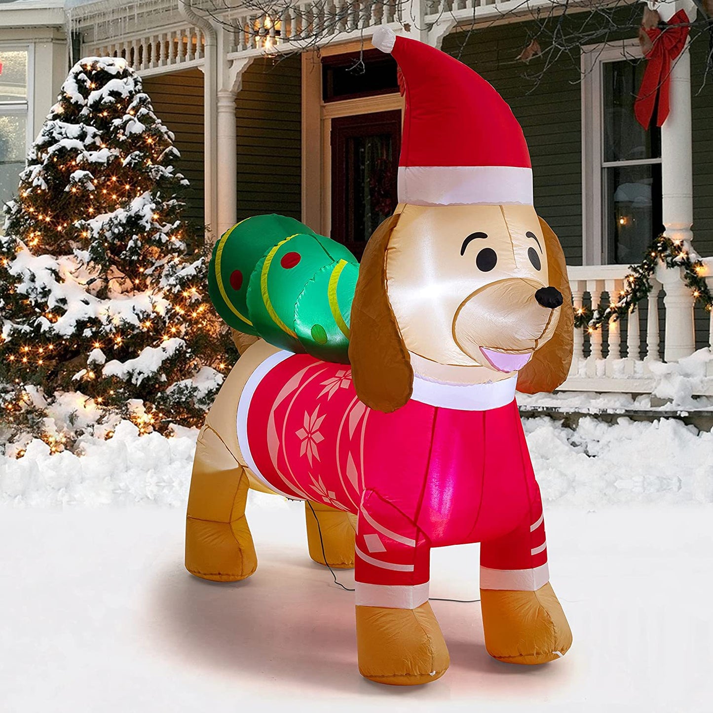 Inflatable dog in a Santa outfit with a Christmas tree on its back, placed in a snowy yard beside a decorated house and lit Christmas tree.