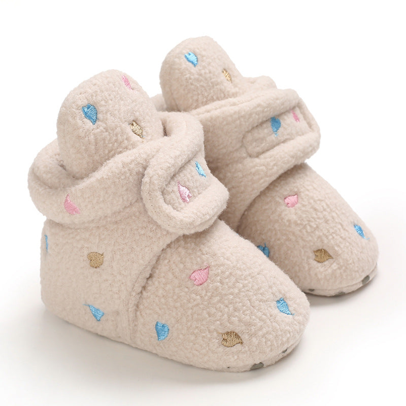 Cream-colored baby booties with a soft, textured finish. Decorated with pink, blue, and gold embroidered hearts. Velcro straps ensure a secure fit for tiny feet.