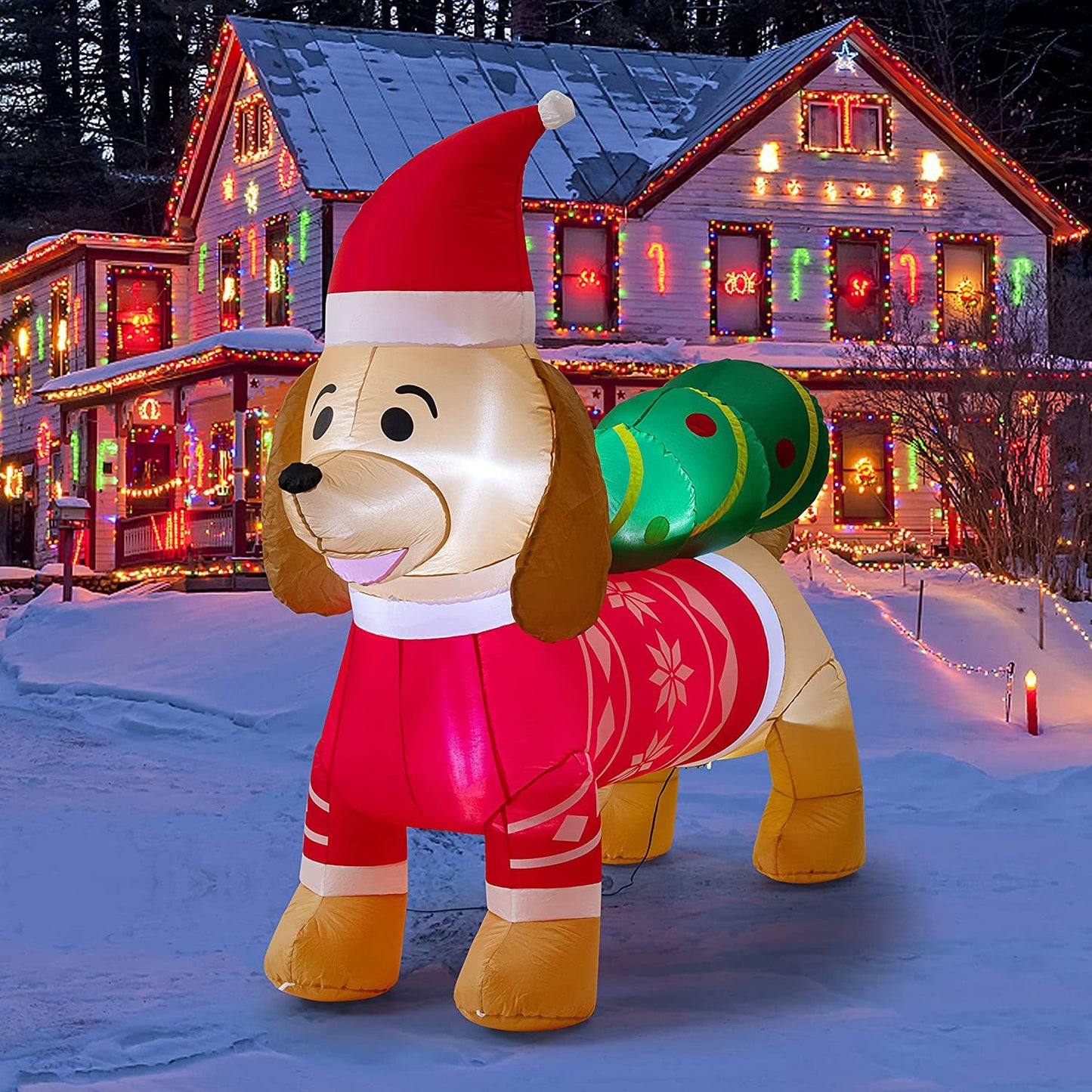 Inflatable dog in a Santa hat and sweater with Christmas designs, standing in front of a festive house adorned with colorful holiday lights in a snowy setting.