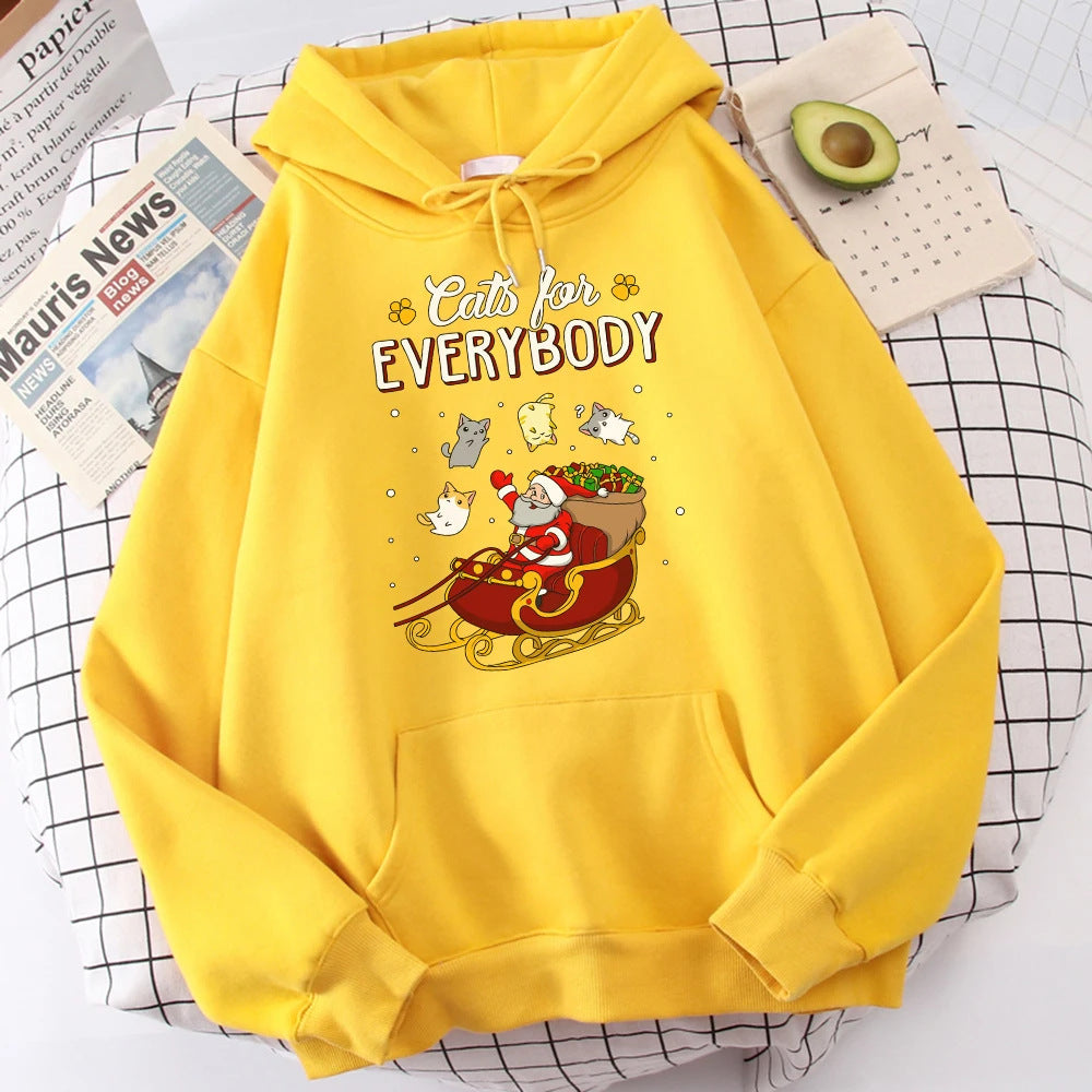 Bright yellow hoodie featuring Santa in a sleigh with cats and gifts. The text reads "Cats for Everybody." Set on a checkered cloth with an avocado and a newspaper nearby.
