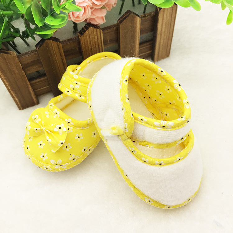 Yellow and white baby shoes with daisy patterns, featuring a small bow on one shoe, placed on a soft surface with a wooden fence and pink flowers in the background.