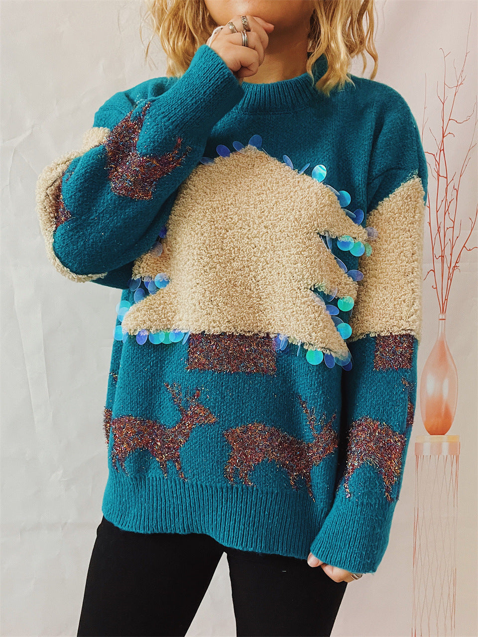 Person wearing a teal sweater with textured beige tree and sequins, adorned with glittery reindeer patterns. Background includes a minimalistic vase with branches.