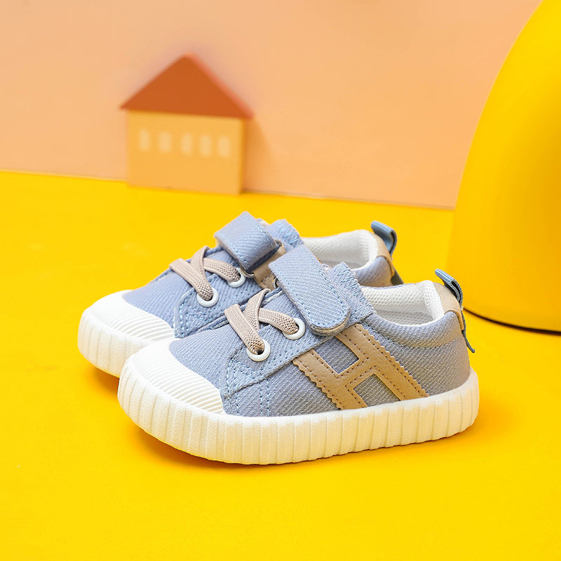 Light blue toddler sneakers with Velcro straps, beige laces, and white soles on a yellow backdrop. A small house decoration is in the background.