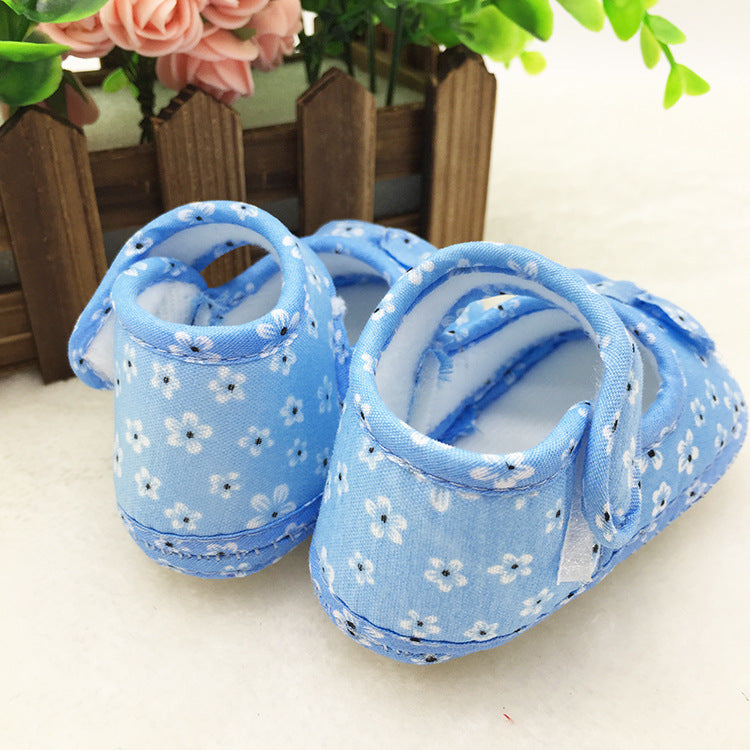Blue baby shoes with white floral pattern are placed near a wooden fence. Pink and green artificial flowers are visible in the background.