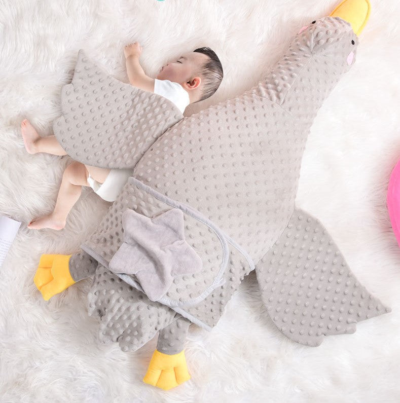 A baby sleeps peacefully on a fluffy white rug, cuddled next to a large plush goose with gray, textured fabric and yellow feet, providing comfort with its wing draped over the child.