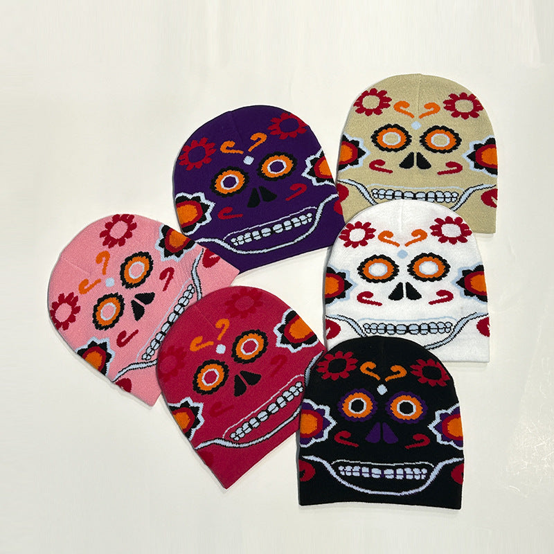 Six knitted beanies with colorful Day of the Dead-style skull designs in purple, beige, pink, red, white, and black are arranged in a circular pattern on a light background.