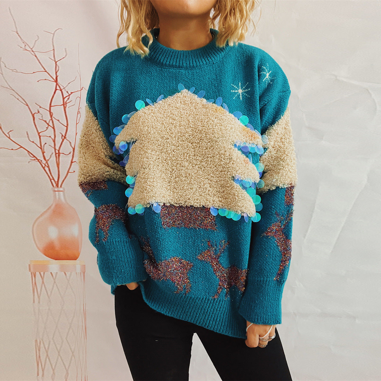 A person wearing a teal sweater with a textured beige Christmas tree design and sequin accents. Reindeer patterns decorate the sleeves. A vase with bare branches stands nearby.