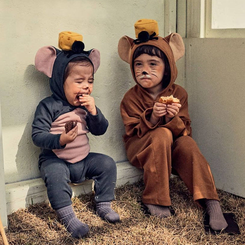 Two toddlers in mouse costumes with cheese hats sit against a wall. The toddler in gray cries while holding food; the one in brown looks on, holding a sandwich, both seated on grass.