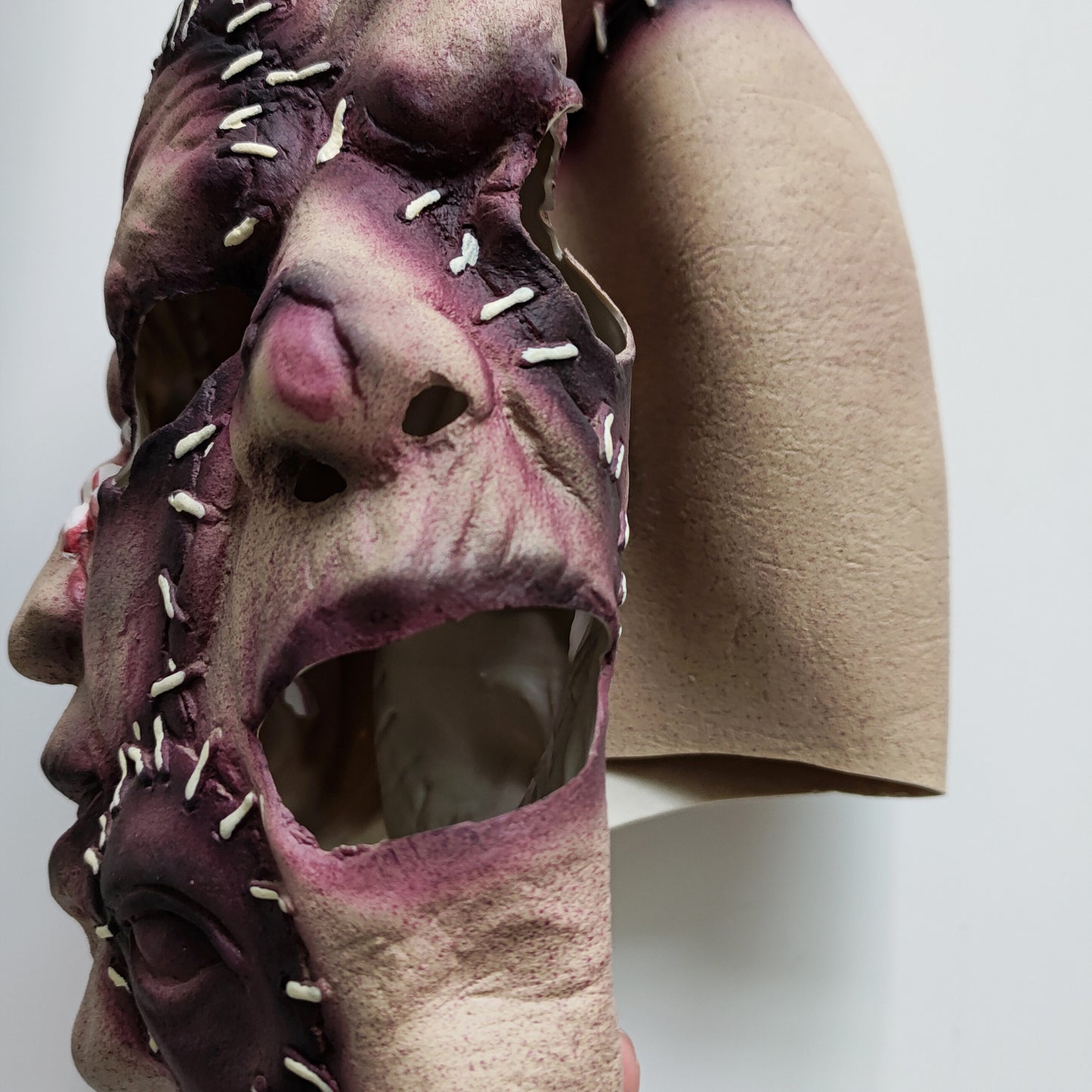 Close-up of a textured, multicolored mask with multiple faces, featuring stitched seams and open eye holes. The mask is attached to a beige, fabric-like material.