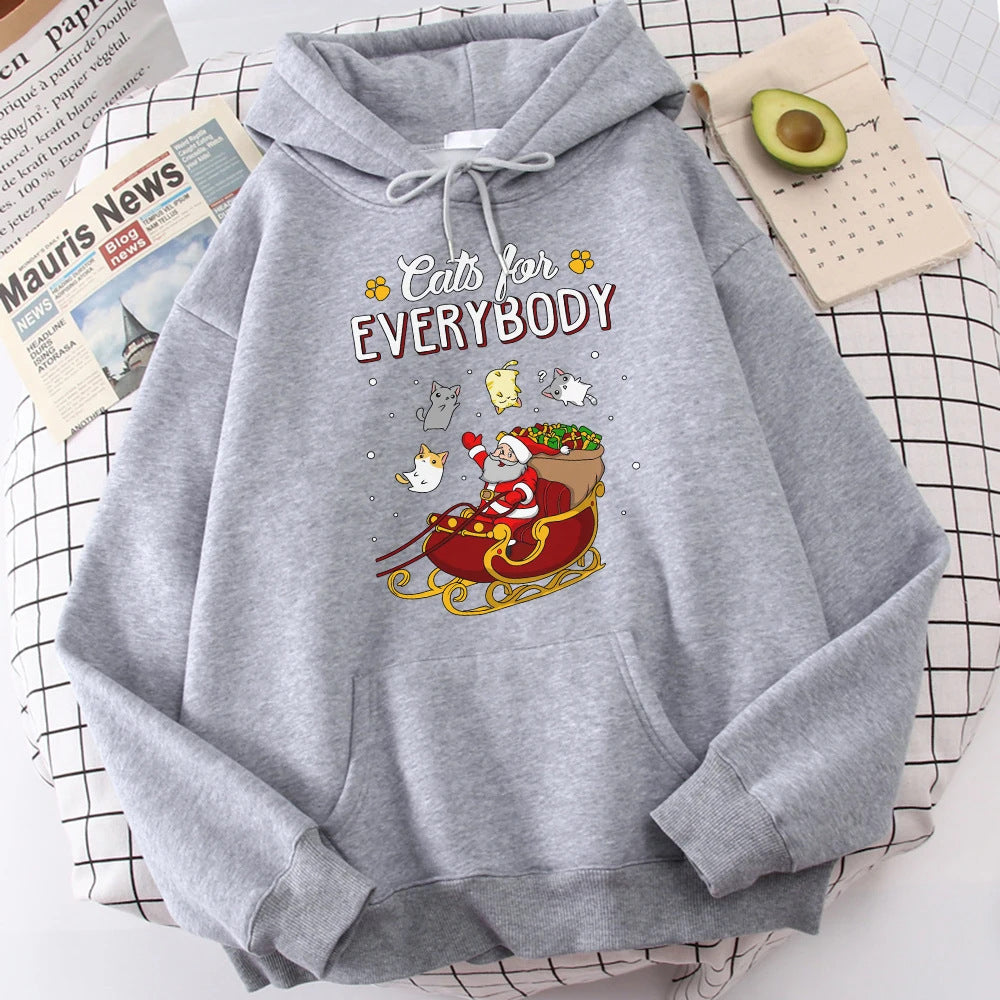 Gray hoodie with "Cats for Everybody" text and a Santa cat in a sleigh with cheerful cats. Laid on a grid-pattern surface beside newspapers and half an avocado.