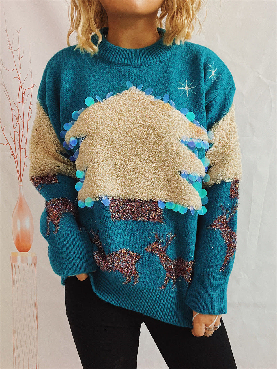 A person wearing a teal sweater with a fluffy beige Christmas tree design, decorated with shiny sequins and reindeer motifs. They stand near a decorative tall vase with twigs.