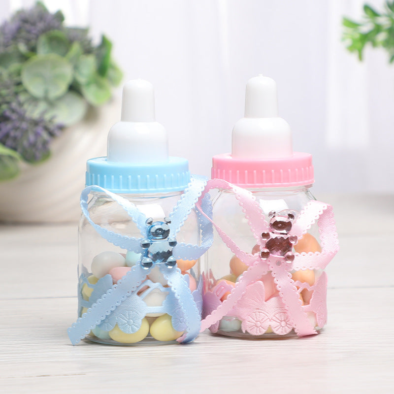 Two transparent baby bottles with blue and pink lids, decorated with matching ribbons and bear charms, filled with colorful candies. Greenery in the blurred background.