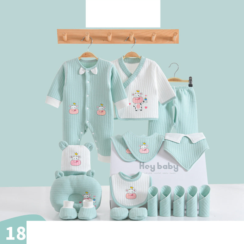 A pastel green and white baby clothing set on a wooden rack includes a jumpsuit, shirts, pants, bibs, mittens, a hat, and a pillow, featuring cute cow designs.