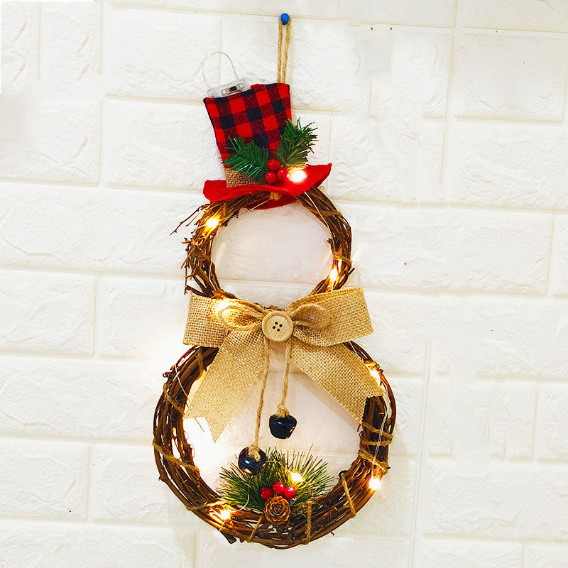 A decorative snowman wreath made of twigs, adorned with warm lights, a red plaid top hat, burlap bow, pinecones, berries, and a pine sprig, hangs on a textured white wall.