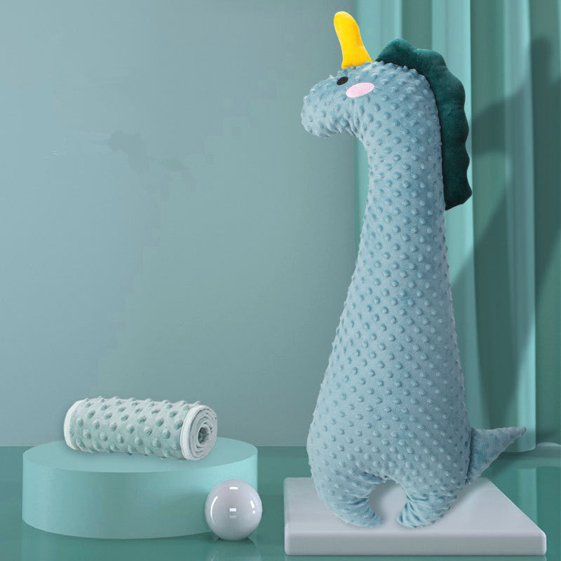 Blue plush dinosaur with a yellow horn and green spikes, standing on a white platform against a teal background. A rolled patterned cloth and white ball are on a nearby pedestal.