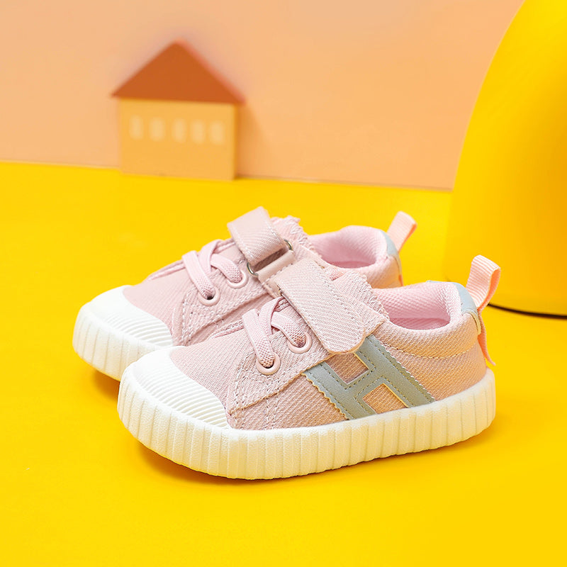 Pink toddler sneakers with velcro straps and white soles on a yellow surface with a small house decoration in the background.