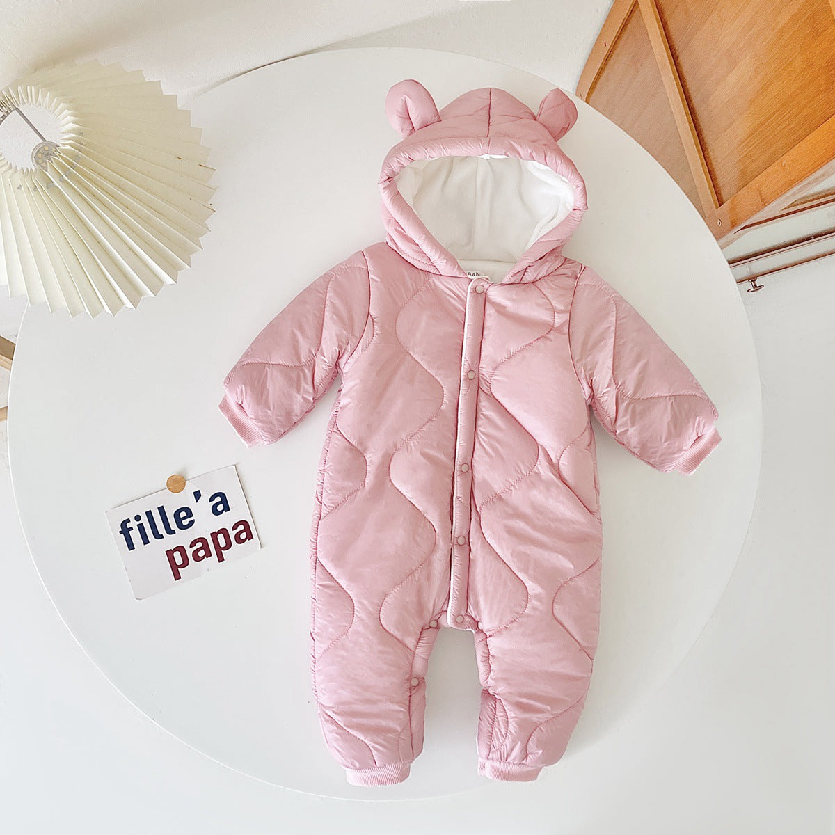 A pink quilted baby snowsuit with bear ears lies on a white surface next to a card with the text "fille'a papa" and a white pleated lampshade.