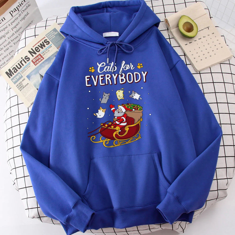 Blue hoodie with "Cats for Everybody" above a Santa graphic riding a sleigh, surrounded by playful cats. Nearby, a halved avocado, calendar, and newspaper sit on a table.