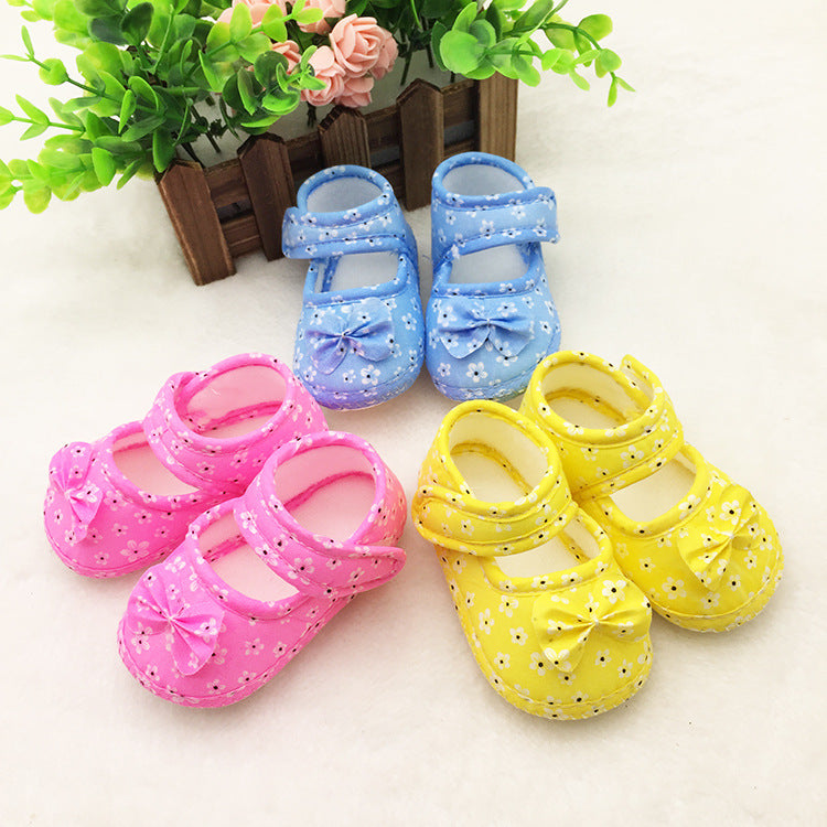 Three pairs of baby shoes: pink, blue, and yellow, each decorated with small white flowers and bows, placed on a light surface near a small plant arrangement.
