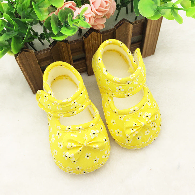 Yellow baby shoes with white floral patterns and bows, placed on a fluffy cream surface. A wooden picket fence with pink and green artificial plants is in the background.