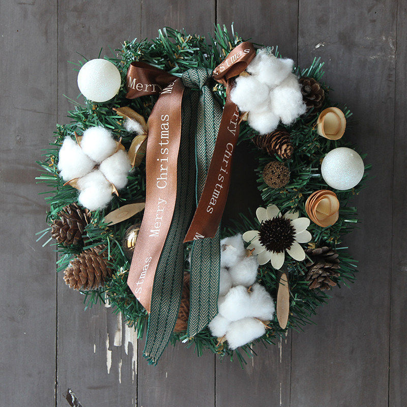 Festive Christmas wreath with cotton, pinecones, white flowers, and gold accents on dark wood. Brown ribbon reads "Merry Christmas" alongside green chevron-patterned ribbon.