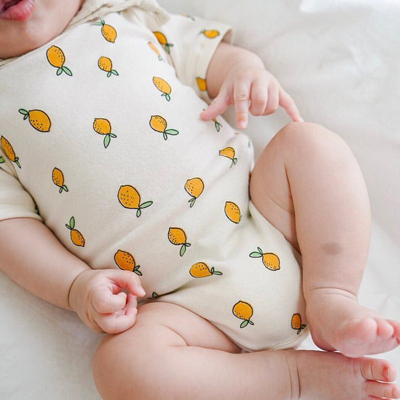 Baby wearing a white onesie patterned with small, yellow lemons, lying on a soft surface. The baby's legs and arms are bent, showcasing the playful lemon design.