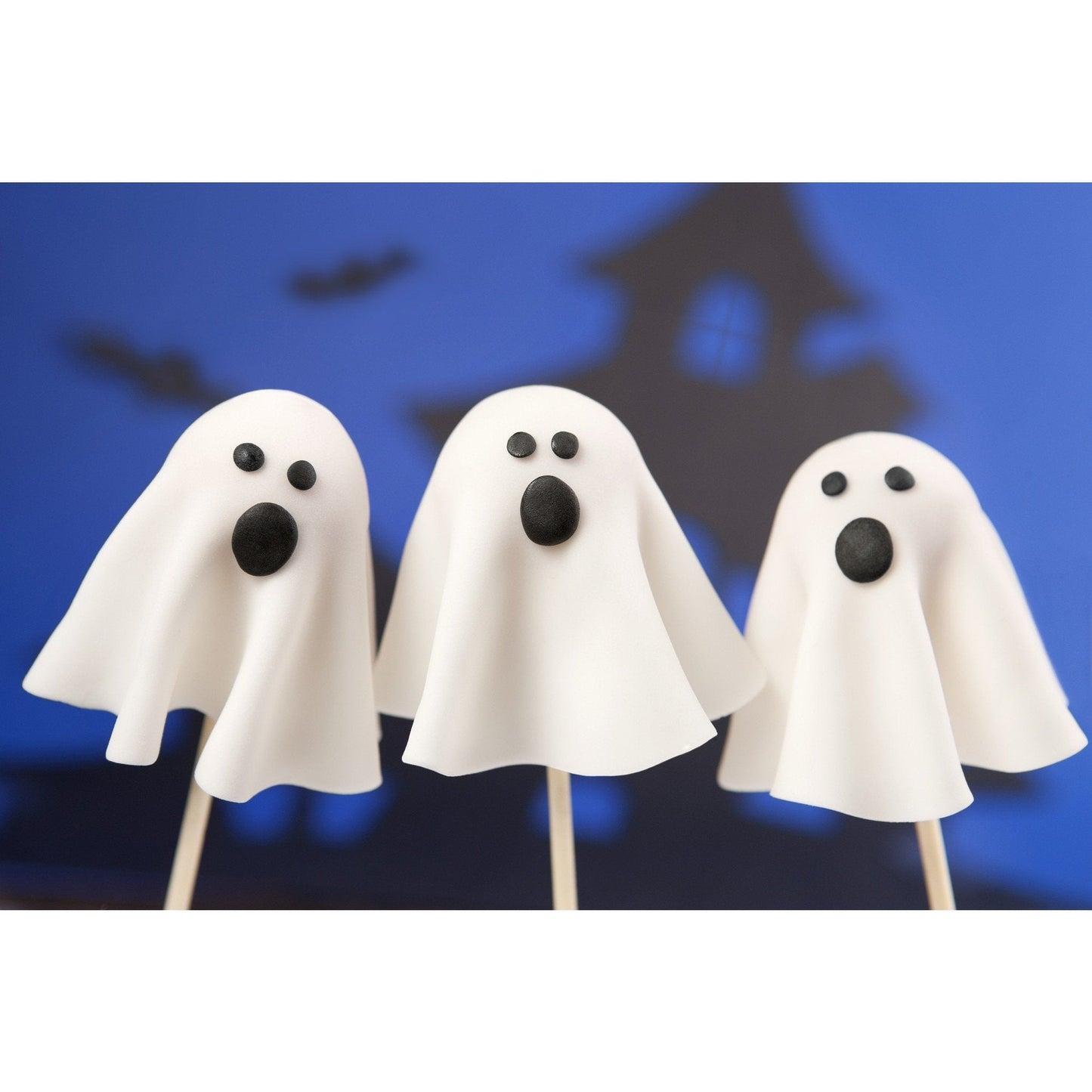 Three ghost-shaped cake pops with white cloth-like coverings and black eyes and mouths against a blue Halloween-themed background with bat and haunted house silhouettes.