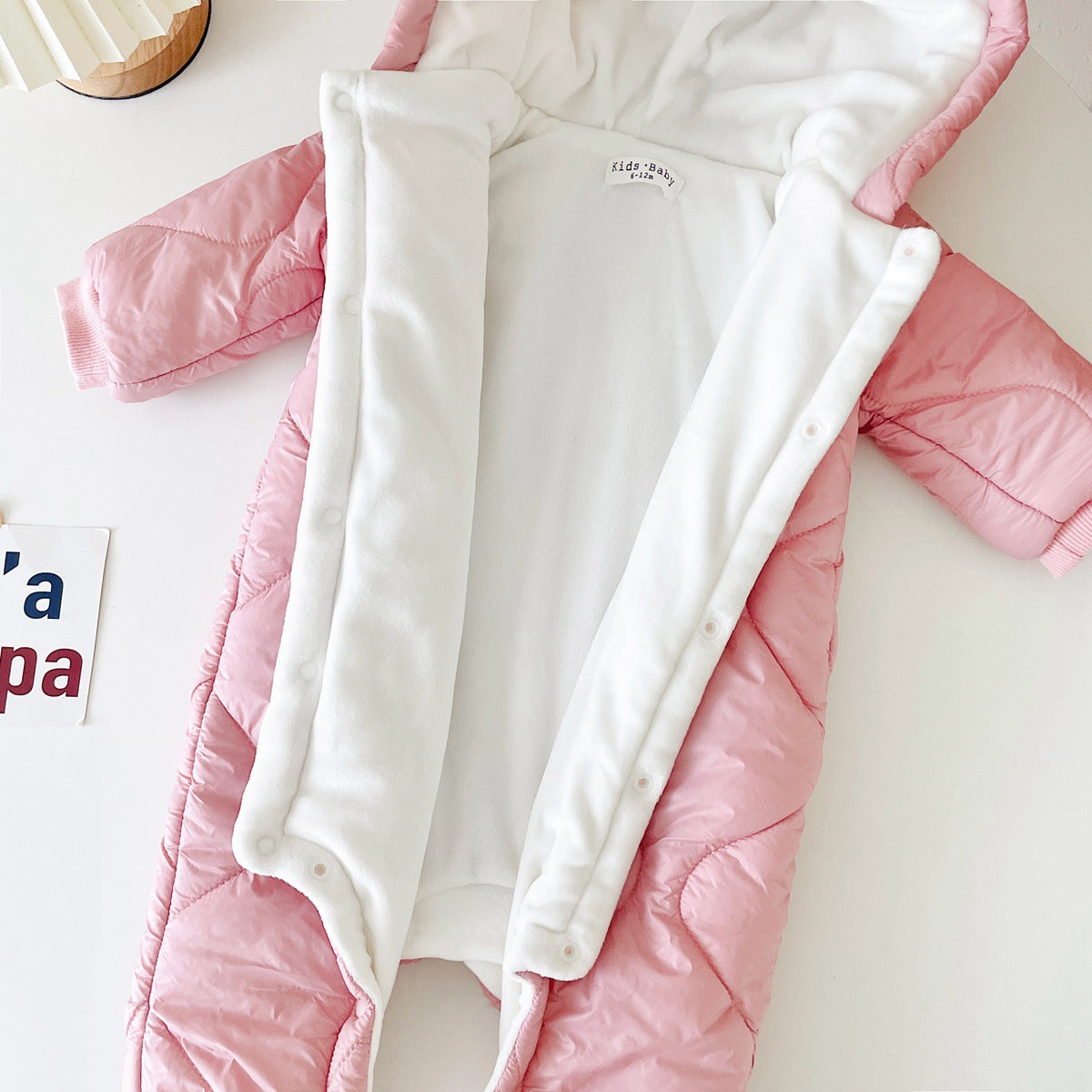 Pink quilted baby snowsuit with a white fleece lining, open to reveal interior. It has a hood, sleeves, and button closures. Nearby, a partial view of a sign with letters.
