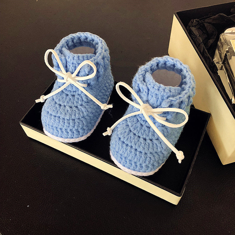 Blue crocheted baby booties with white laces, displayed in a black and cream box on a dark surface.