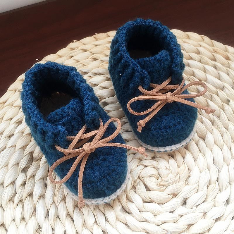 Knitted baby shoes in dark blue with brown laces, placed on a woven circular mat, creating a cozy and handmade feel.