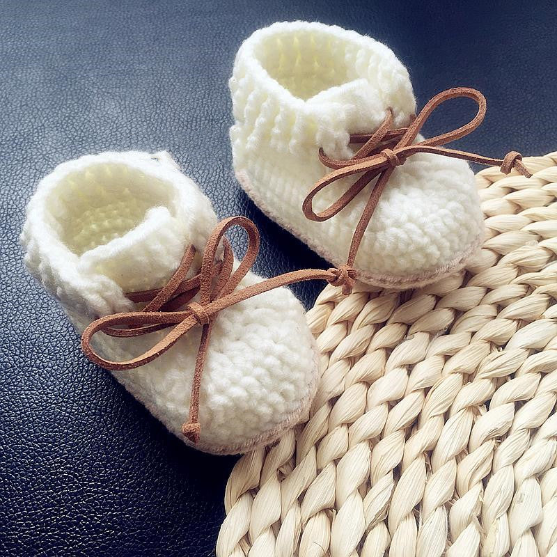 Knitted white baby booties with brown laces on a black leather background, next to a woven straw mat.