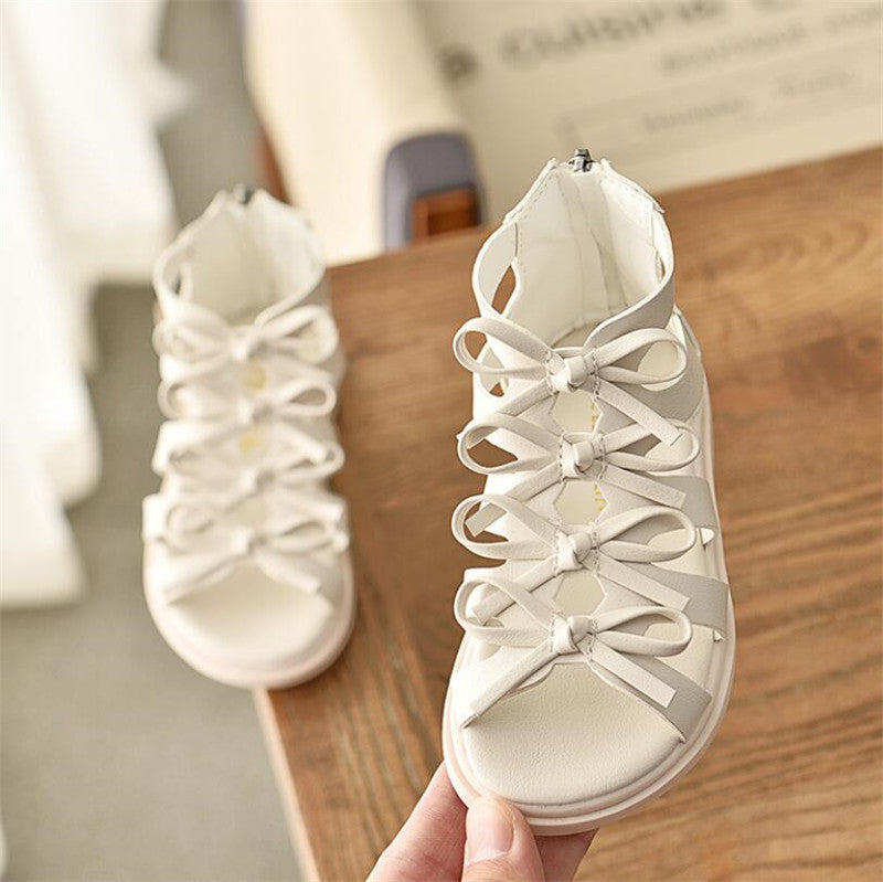 White open-toe sandals with multiple decorative bows along the front, displayed on a wooden surface. One shoe is held up for a closer look. Simple and elegant design.