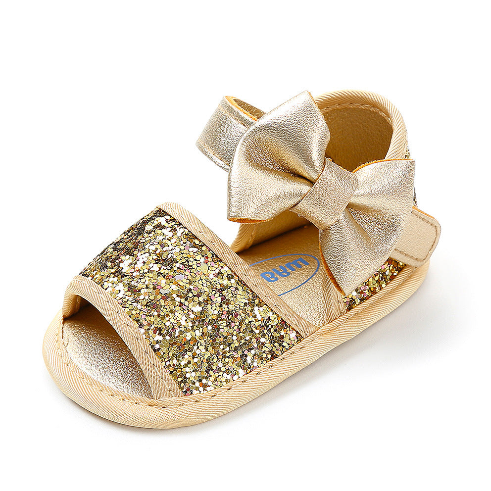 Gold toddler sandal with glittery straps and a metallic bow on the side. The shoe has an open toe design and a soft sole, ideal for infants.