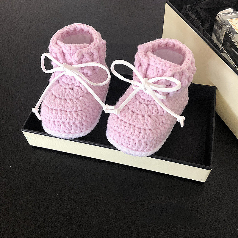 Pink knitted baby booties with white ribbon bows, placed in a black and white box against a dark background.