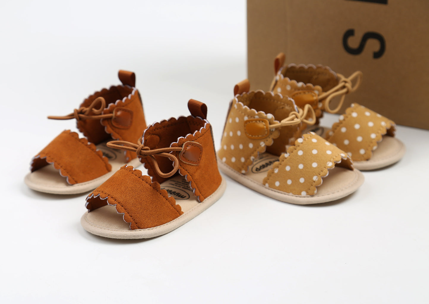 Two pairs of baby sandals on a white surface. One pair is brown with scalloped edges, and the other is light brown with white polka dots, both with laces and a box in the background.