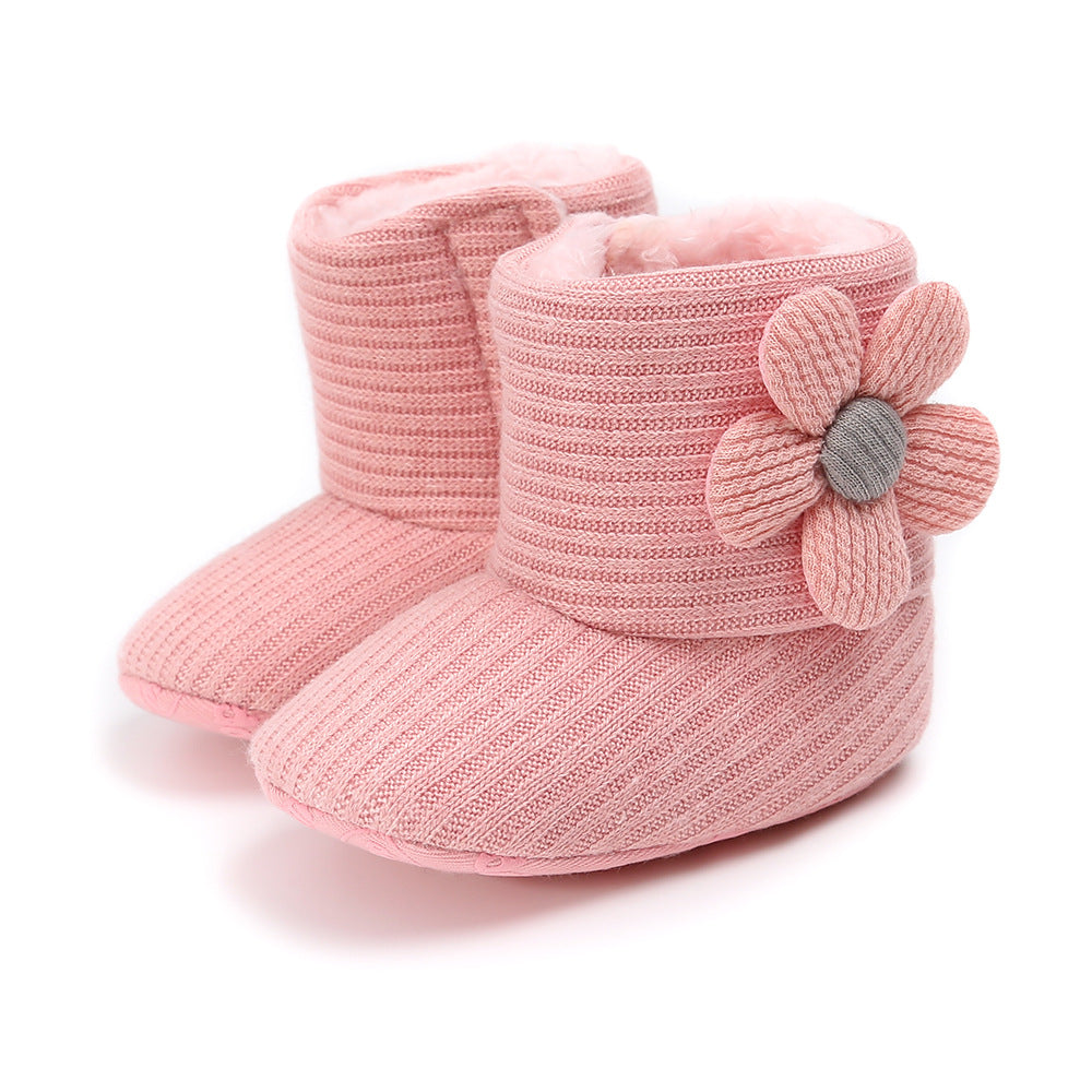 Pink knitted baby booties with ribbed texture and a soft, plush interior. Each bootie features a decorative fabric flower on the side, adding a charming touch.