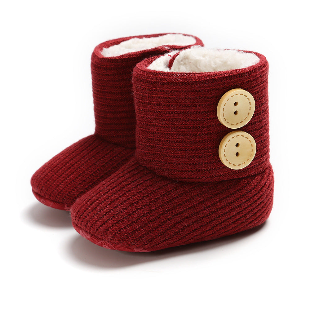 Red knitted baby booties with white fleece lining and two large beige buttons on the side. The texture is ribbed, and they have a soft, cozy appearance.