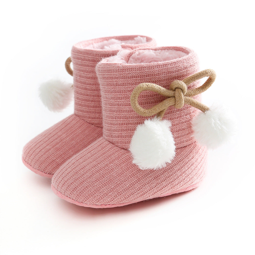 Pink knitted baby booties with a soft, fluffy interior. They feature a bow made of brown cord and white pom-poms on the sides, providing a cute and cozy look.