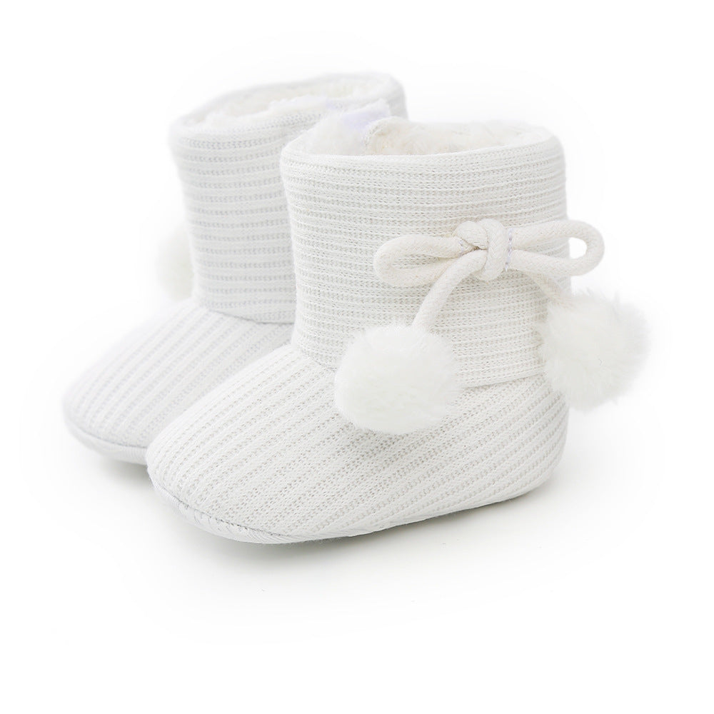 White knitted baby booties with fluffy pom-poms and bow, featuring a soft, ribbed texture. The interior appears plush, providing warmth and comfort.