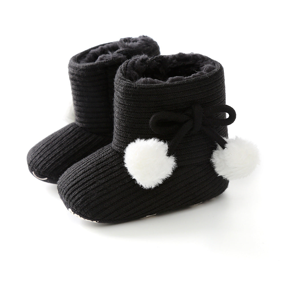 Black knitted baby boots with soft inner lining, adorned with white fluffy pom-poms on the sides and black bows.