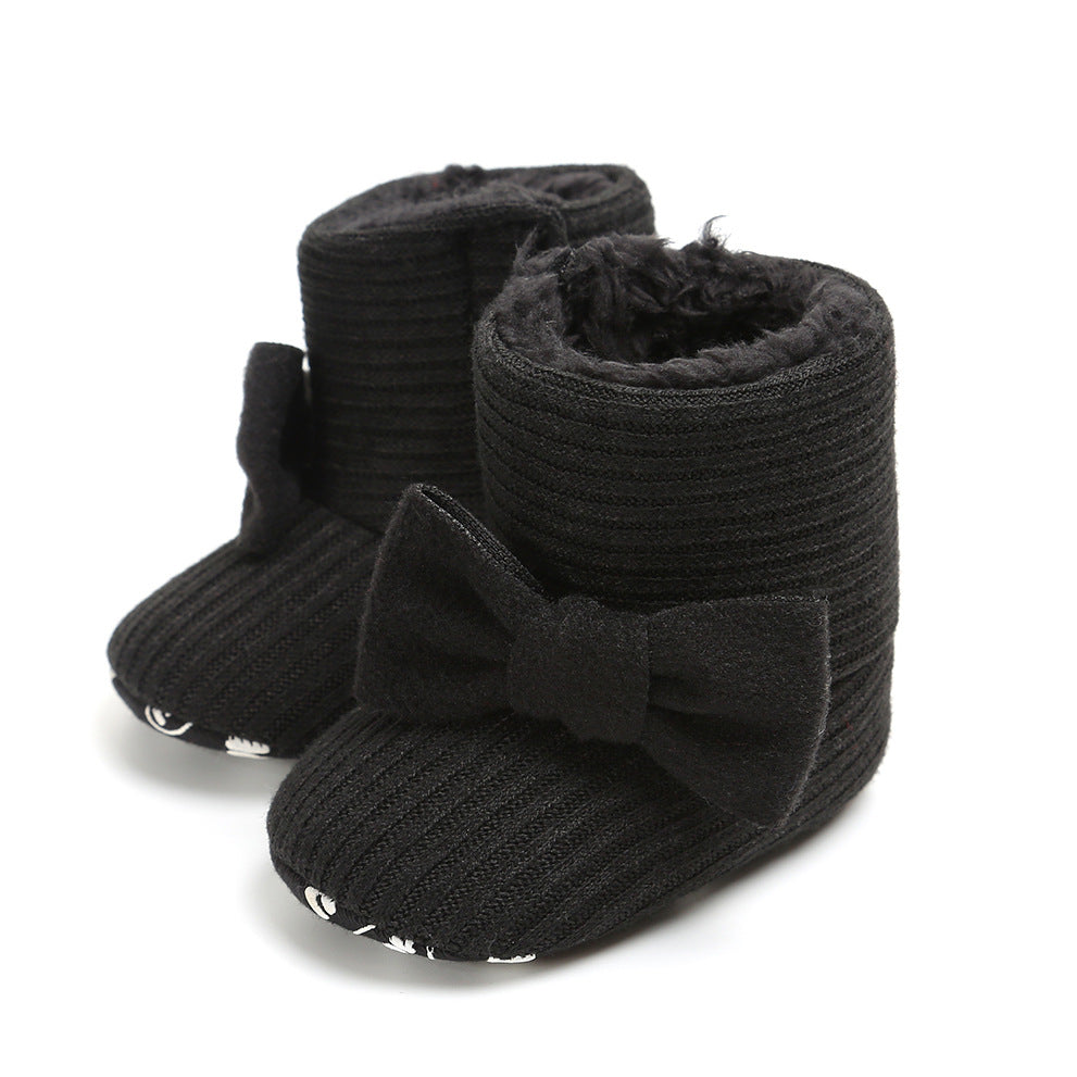 Black knitted baby booties with a plush interior and large front bow, featuring ribbed texture and patterned soles.
