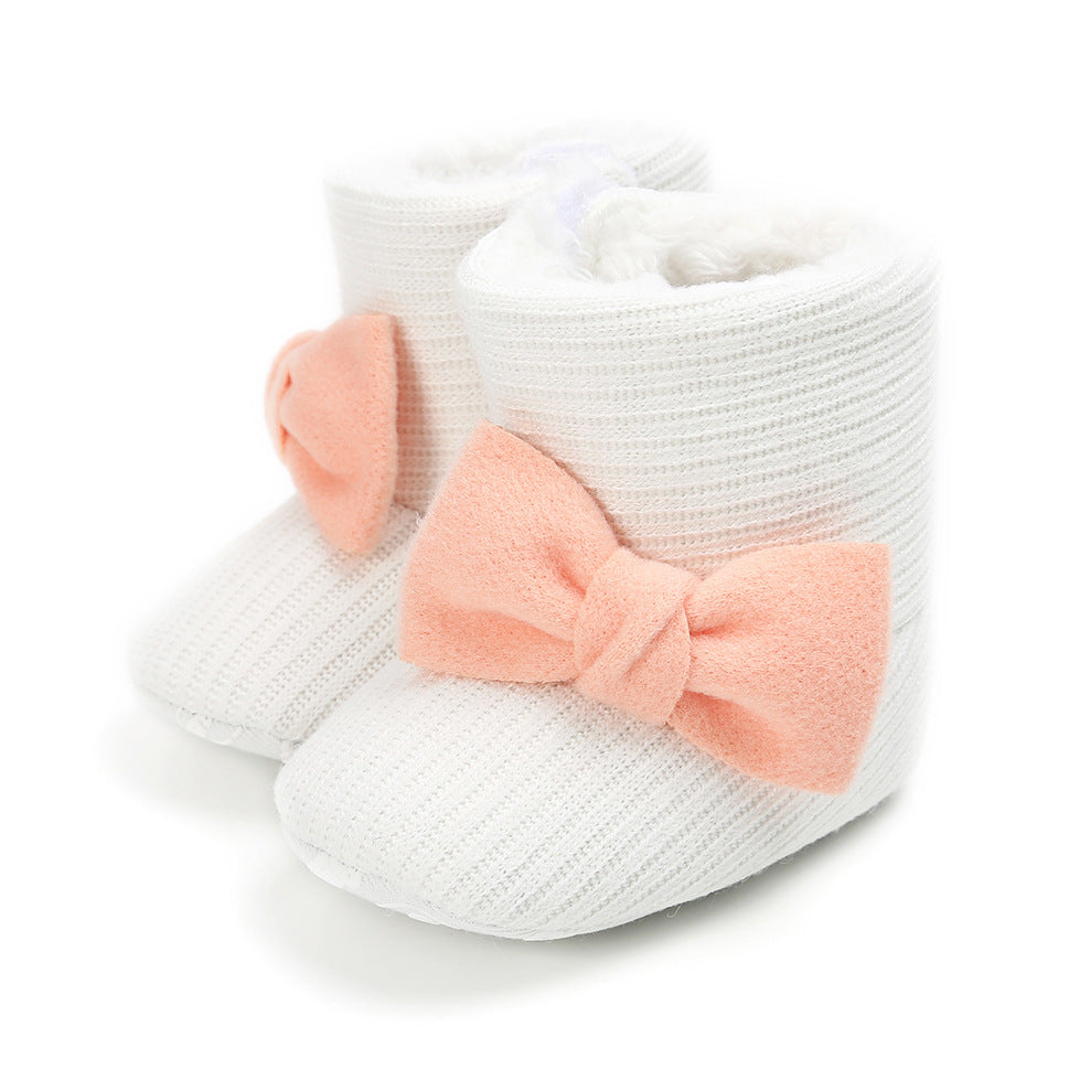 White baby booties with a ribbed texture, adorned with large, soft pink bows on the front. The interior is lined with fluffy white material, adding warmth and comfort.