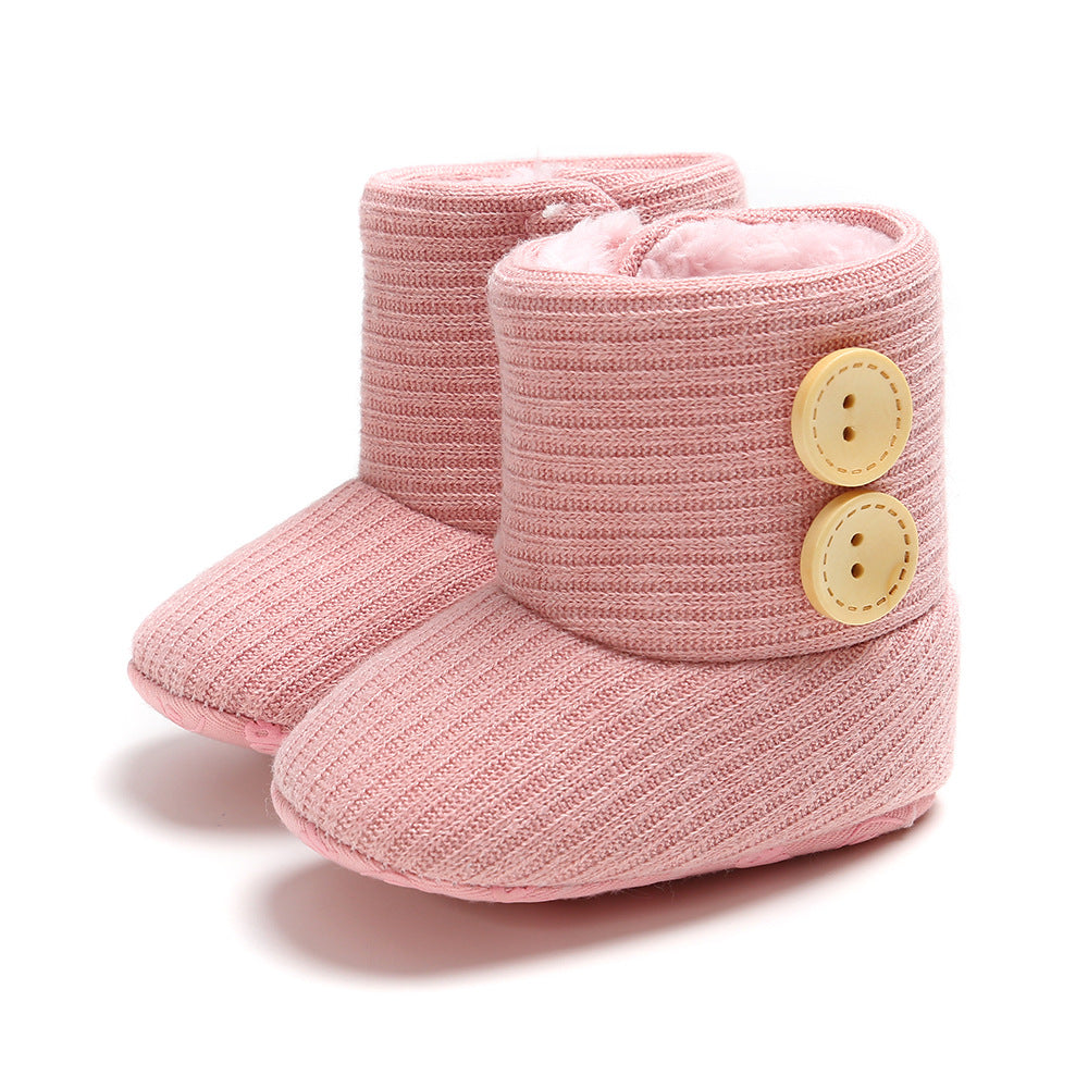 Pink baby booties with a ribbed texture and two large yellow buttons on the sides. The interior appears soft and plush, offering warmth and comfort.