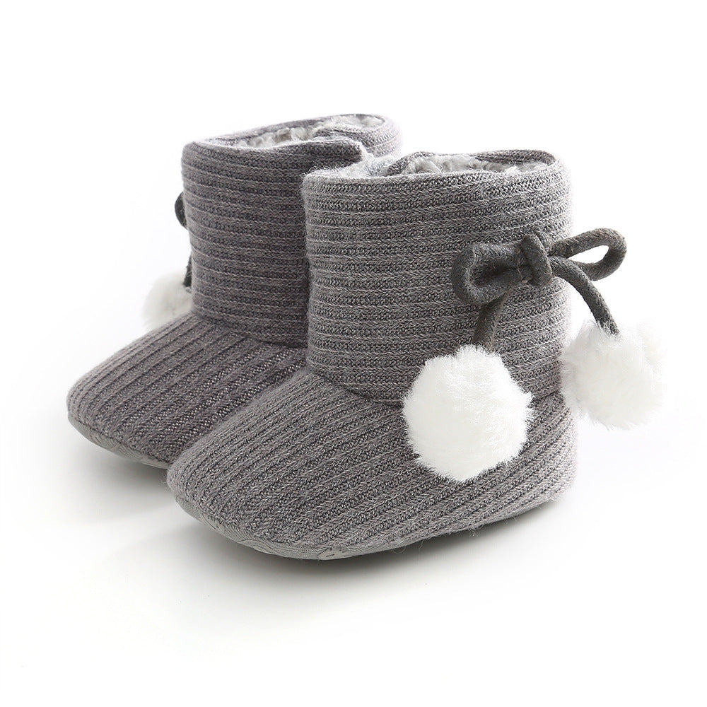 Gray knitted baby booties with ribbed texture, featuring a side bow and fluffy white pom-poms. Soft and cozy, ideal for keeping little feet warm.