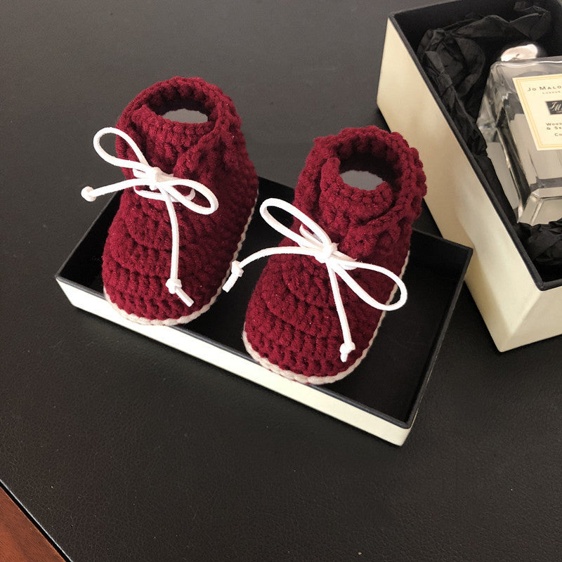 Maroon crocheted baby booties with white laces in a black box, alongside a boxed perfume on a dark surface.