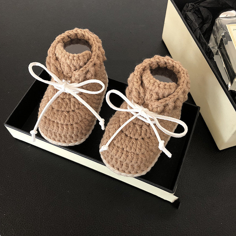 Brown crocheted baby booties with white laces in a black box on a dark surface, next to a gift box with black tissue paper and a perfume bottle.