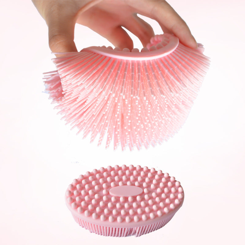 A hand holds a flexible, pink silicone scrubber with bristles, above a flat round base with raised bumps, designed for cleaning or exfoliating.
